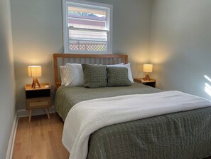 Une chambre à coucher avec une tête de lit en bois, deux tables de chevet avec des lampes, un lit recouvert d’une courtepointe verte matelassée et une fenêtre avec des stores.