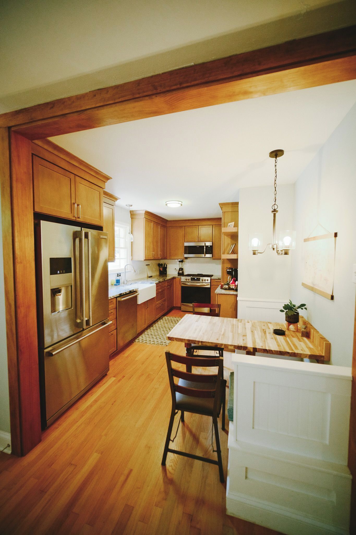 Una cocina con armarios de madera, electrodomésticos de acero inoxidable, una zona de comedor con mesa y sillas, y una lámpara colgante.