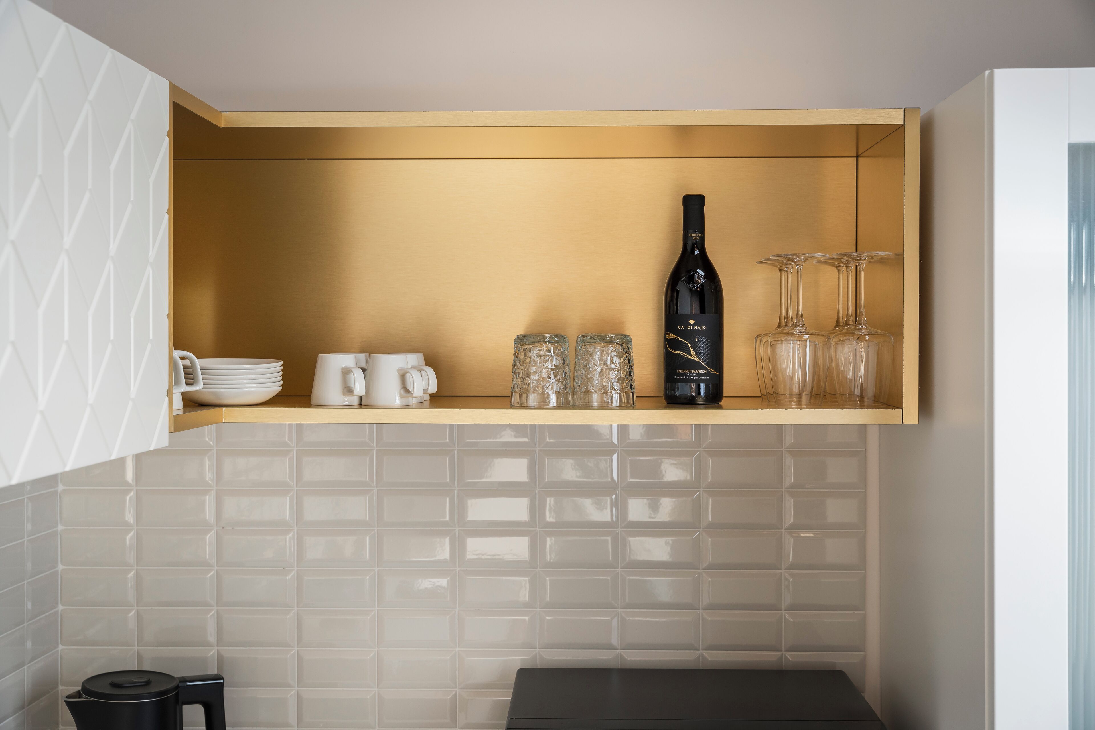 A kitchen with a built-in shelf holding a wine bottle, glasses, and white mugs.