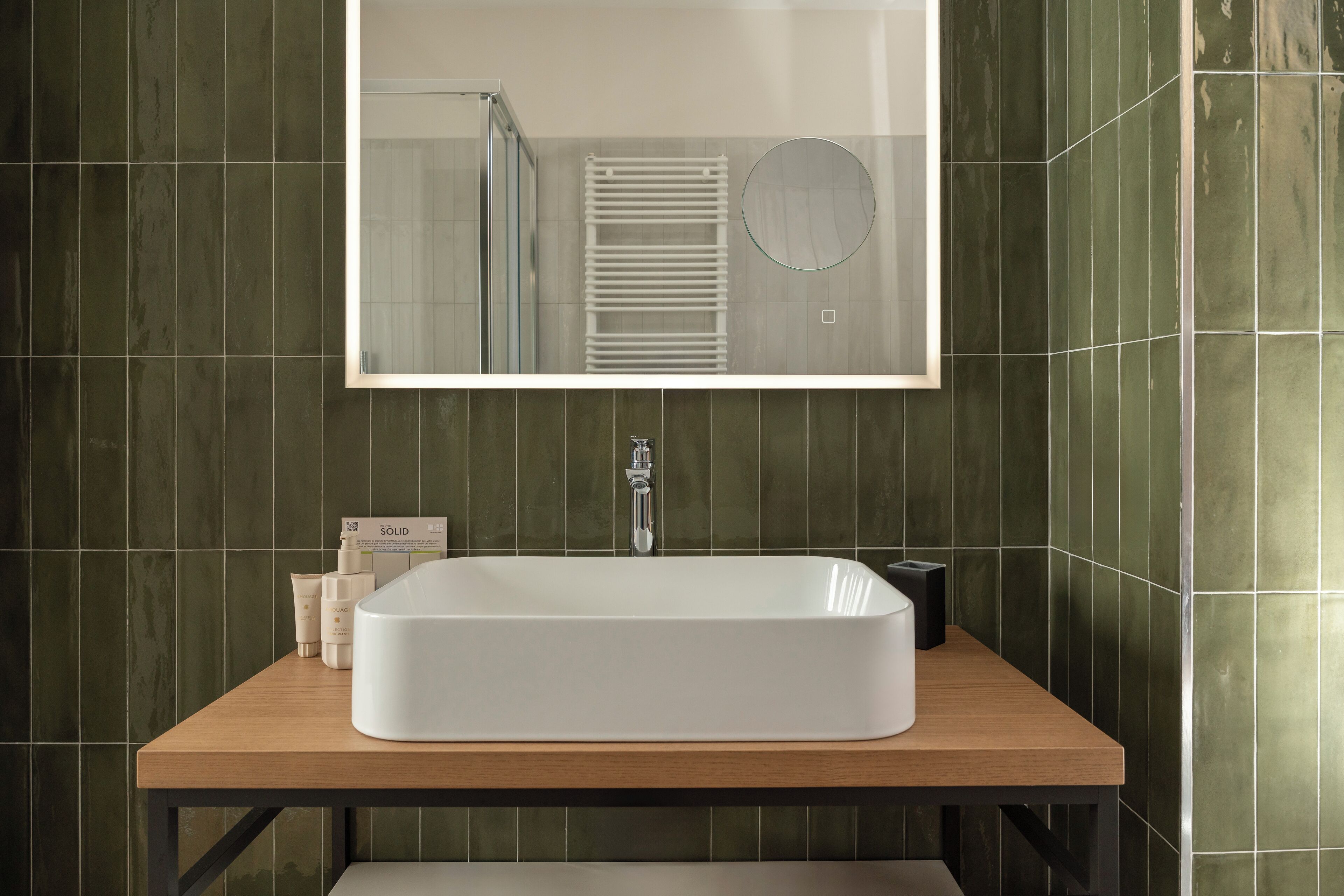 Une salle de bain avec un lavabo rectangulaire blanc, un miroir au-dessus et des murs recouverts de carreaux verts.