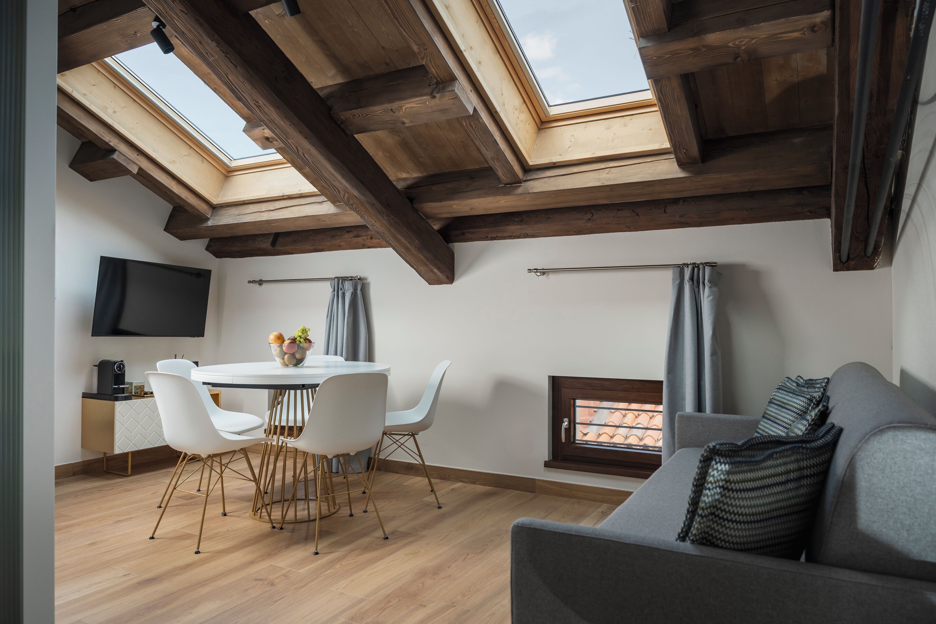 A modern loft with exposed wooden beams, a skylight, a dining area with white chairs, a gray sofa, and a flat-screen TV mounted on the wall.