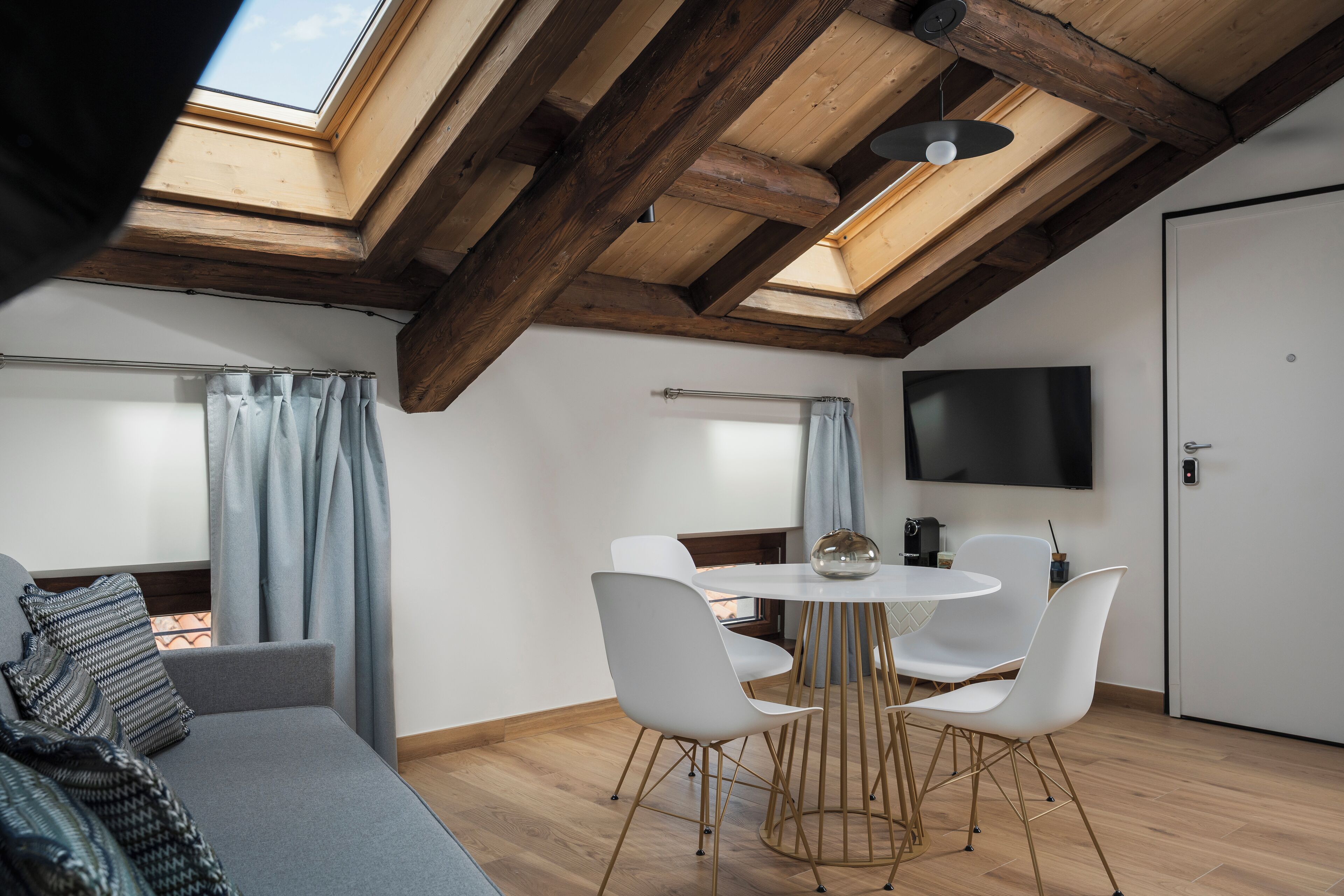 A modern loft bedroom with a skylight, a dining area with white chairs, and a flat-screen TV.
