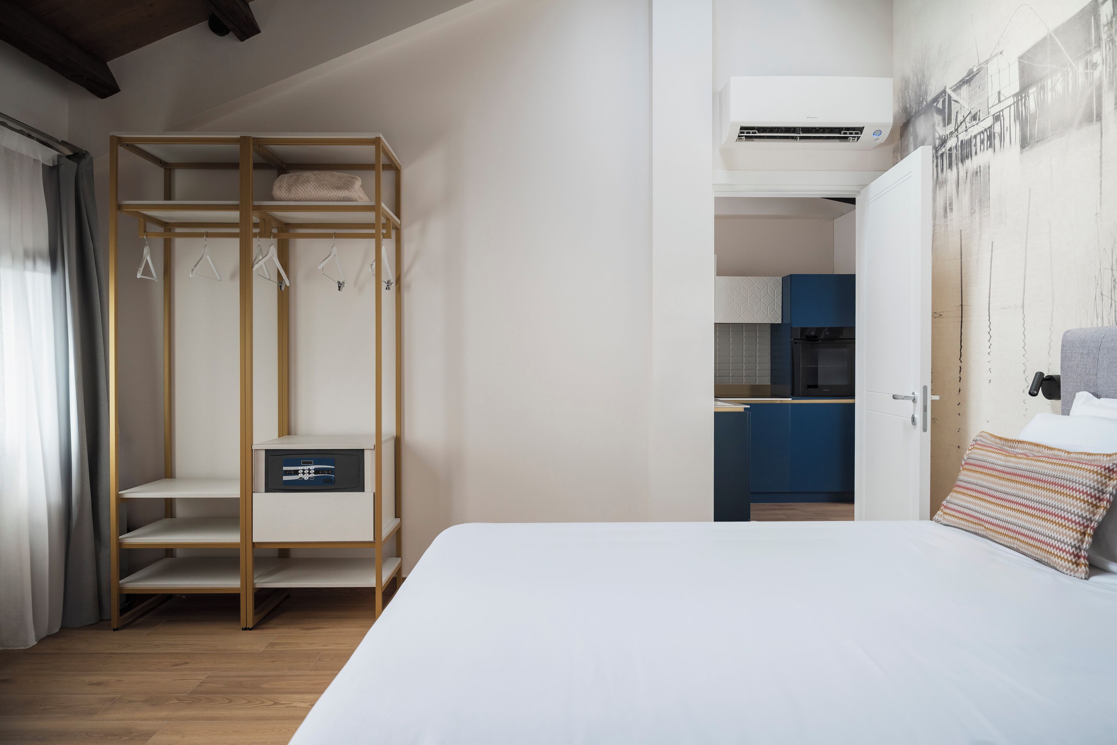 A bedroom with a bed, a wardrobe with hanging clothes, a small cabinet, and a kitchenette in the background.