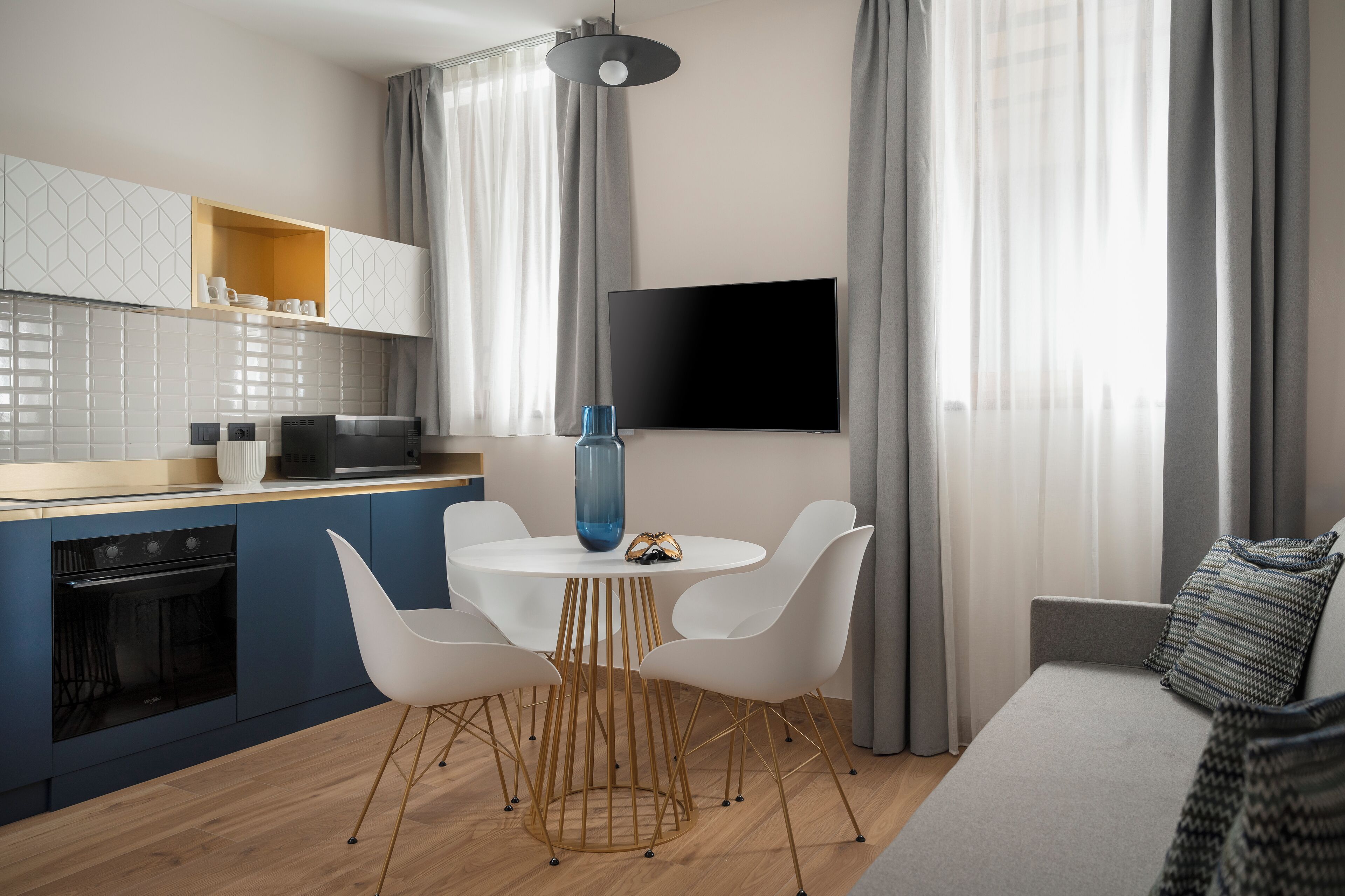 A modern kitchen with a built-in oven, a dining area with a round table and white chairs, and a flat-screen TV mounted on the wall.
