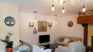 A living room with a TV, two sofas, a coffee table, and a dining table with chairs. There are wall decorations, a clock, and a fireplace. - FALE TAHI - Les Petites rives de la Motte (PLEUGUENEUC)
