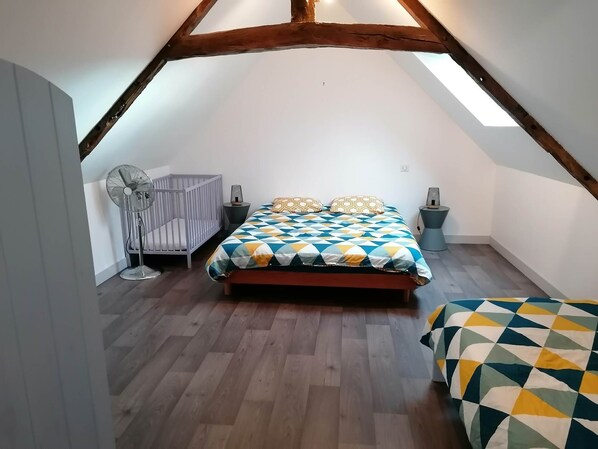 A bedroom with a sloped ceiling, a bed with a patterned bedspread, a crib, a standing fan, and bedside lamps.