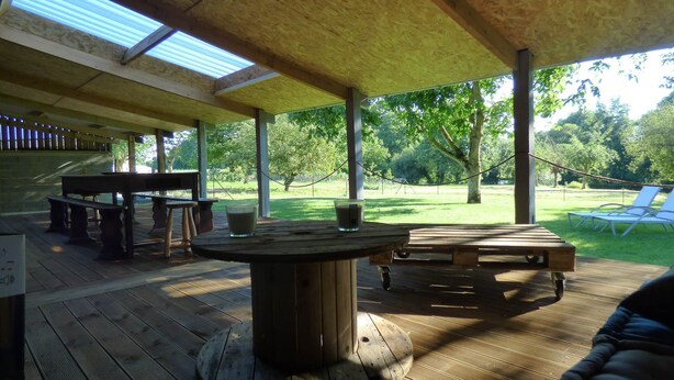 An outdoor covered patio area with wooden tables and benches, a large central table, and a view of a grassy park with trees and a swing set. - FALE LUA - Les Petites rives de la Motte (PLEUGUENEUC)
