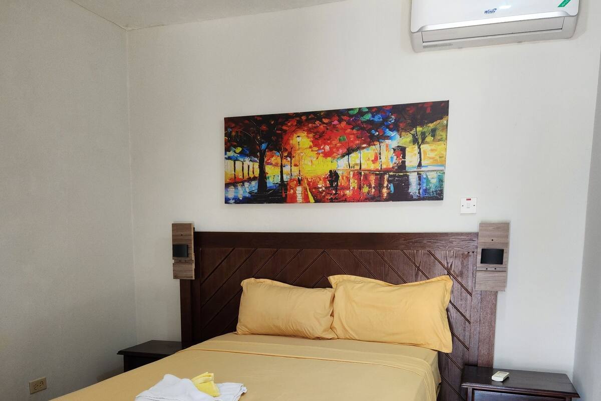 Een hotelkamer met een bed, een schilderij aan de muur, een airconditioner en een klein nachtkastje.