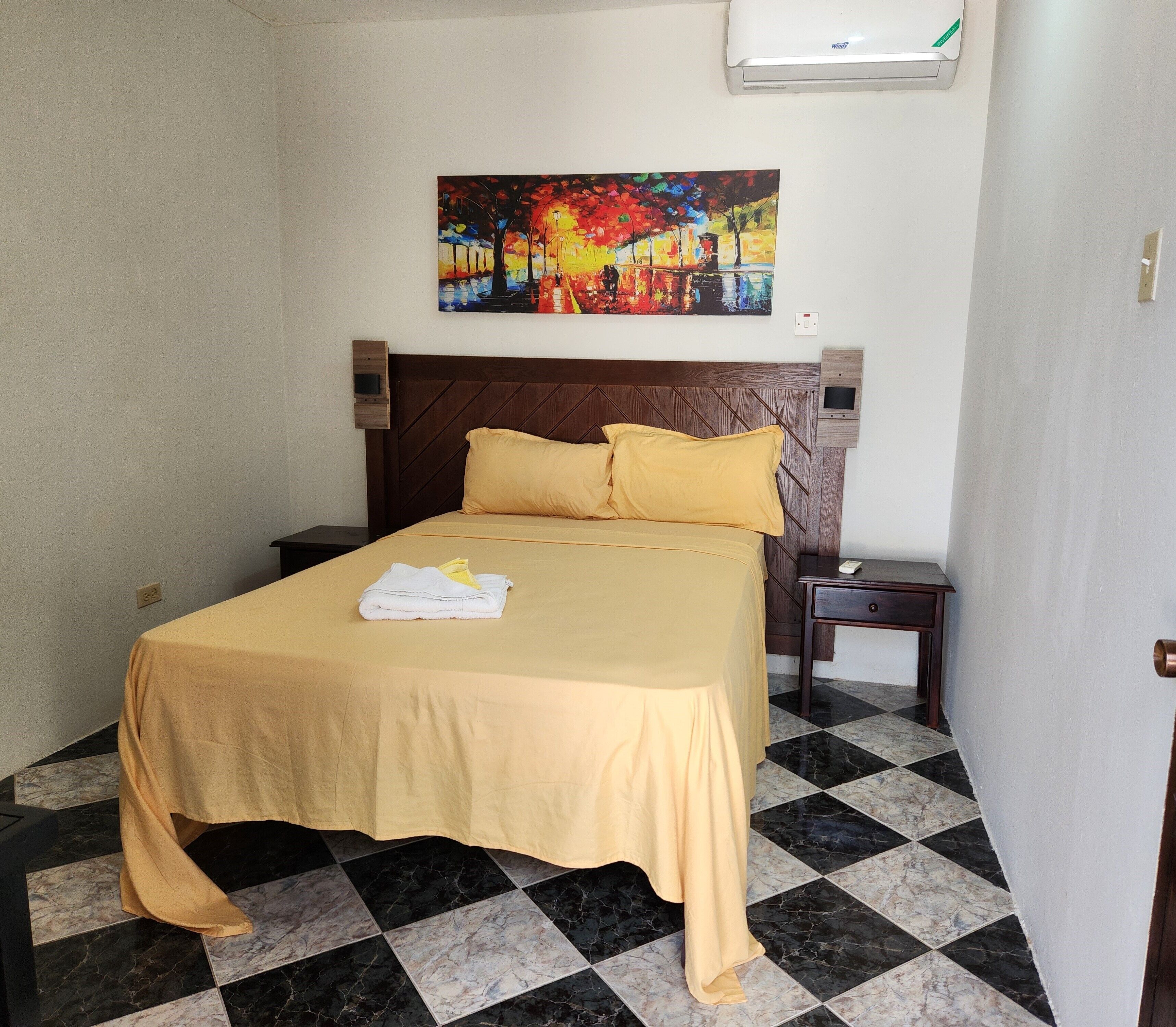 Una habitación de hotel con cama, un cuadro sobre la cama, una mesita de noche y un aire acondicionado de pared.