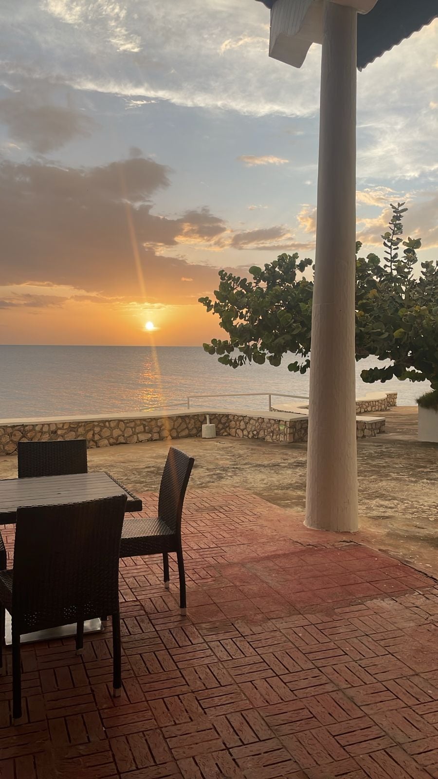 An outdoor seating area with a view of the sunset over the sea, stone walls, and a tree.