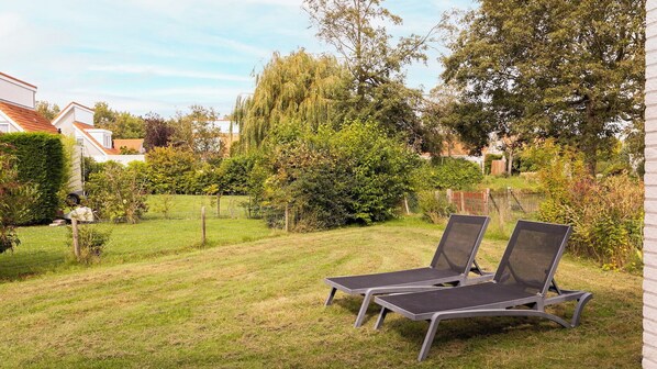 Un jardin avec deux chaises longues, une pelouse verdoyante et une variété d’arbres et d’arbustes.