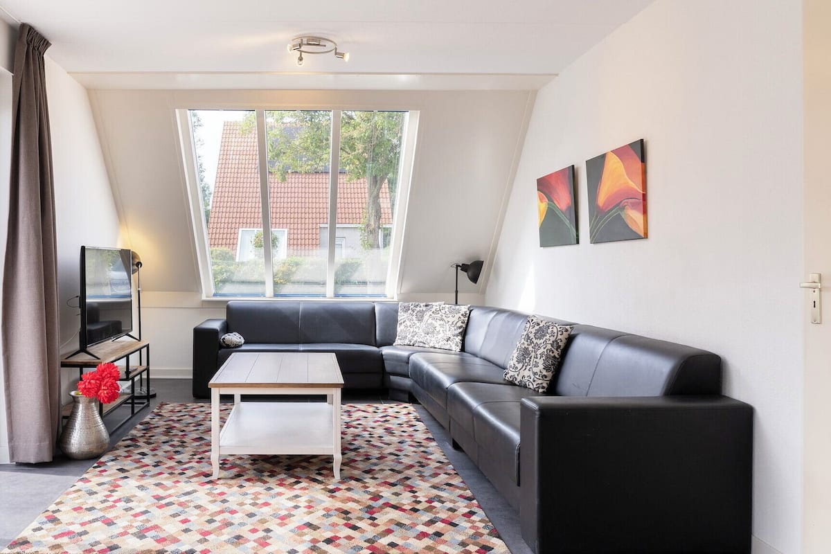 Un salon moderne avec un canapé en cuir noir, une table basse blanche et un tapis à motifs. Une grande fenêtre offre une vue sur un bâtiment aux toits rouges.