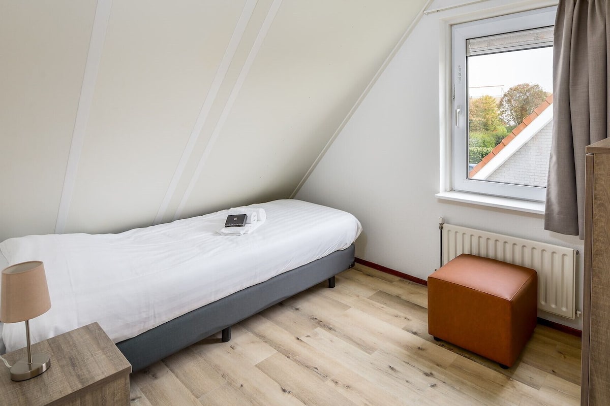 Une petite chambre à coucher, bien rangée, avec un lit simple, une table de chevet avec une lampe, une fenêtre avec des rideaux et un radiateur.