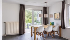 A dining area with a table and chairs, a radiator, curtains, and a view of greenery outside. - Summio Zeeland Village (Scharendijke)