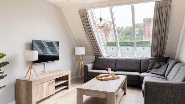 Una sala de estar moderna con televisor de pantalla plana, muebles de madera, un sofá gris y ventanas amplias con cortinas.