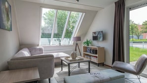 A modern living room with a sofa, armchair, and ottoman, a coffee table, a lamp, a TV on a stand, and a bookshelf. - Summio Zeeland Village (Scharendijke)