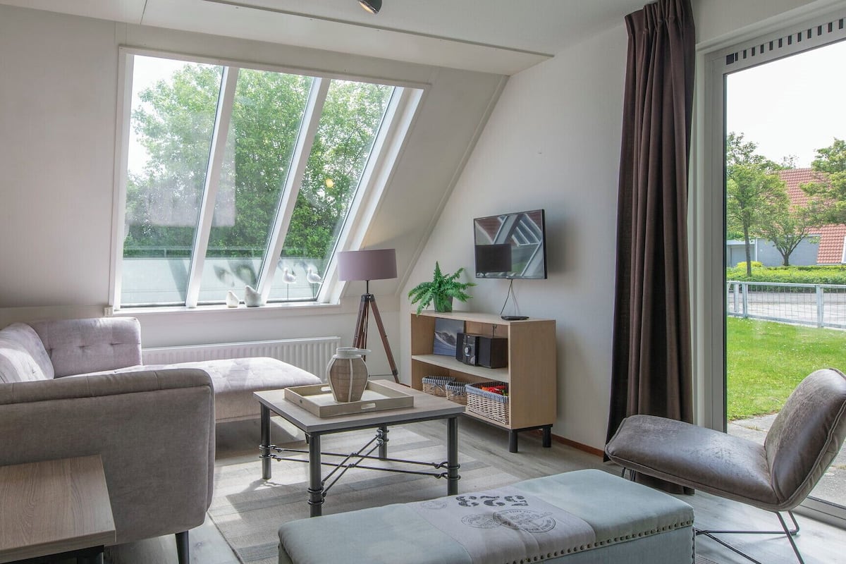 Un salon moderne comprenant un canapé, un fauteuil et un pouf, une table basse, une lampe, un téléviseur posé sur un meuble et une bibliothèque.