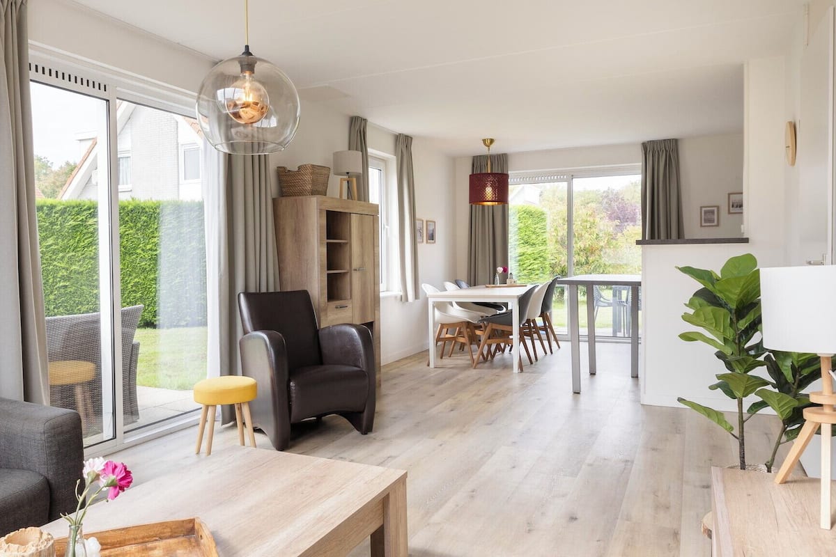 Un salon moderne avec un canapé, une table à manger et une chaise. On y trouve une grande fenêtre avec des rideaux, un meuble en bois et un luminaire suspendu.