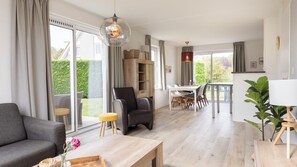 Een moderne woonkamer met een bank, een eettafel en een stoel. Er is een groot raam met gordijnen, een houten kast en een hanglamp.