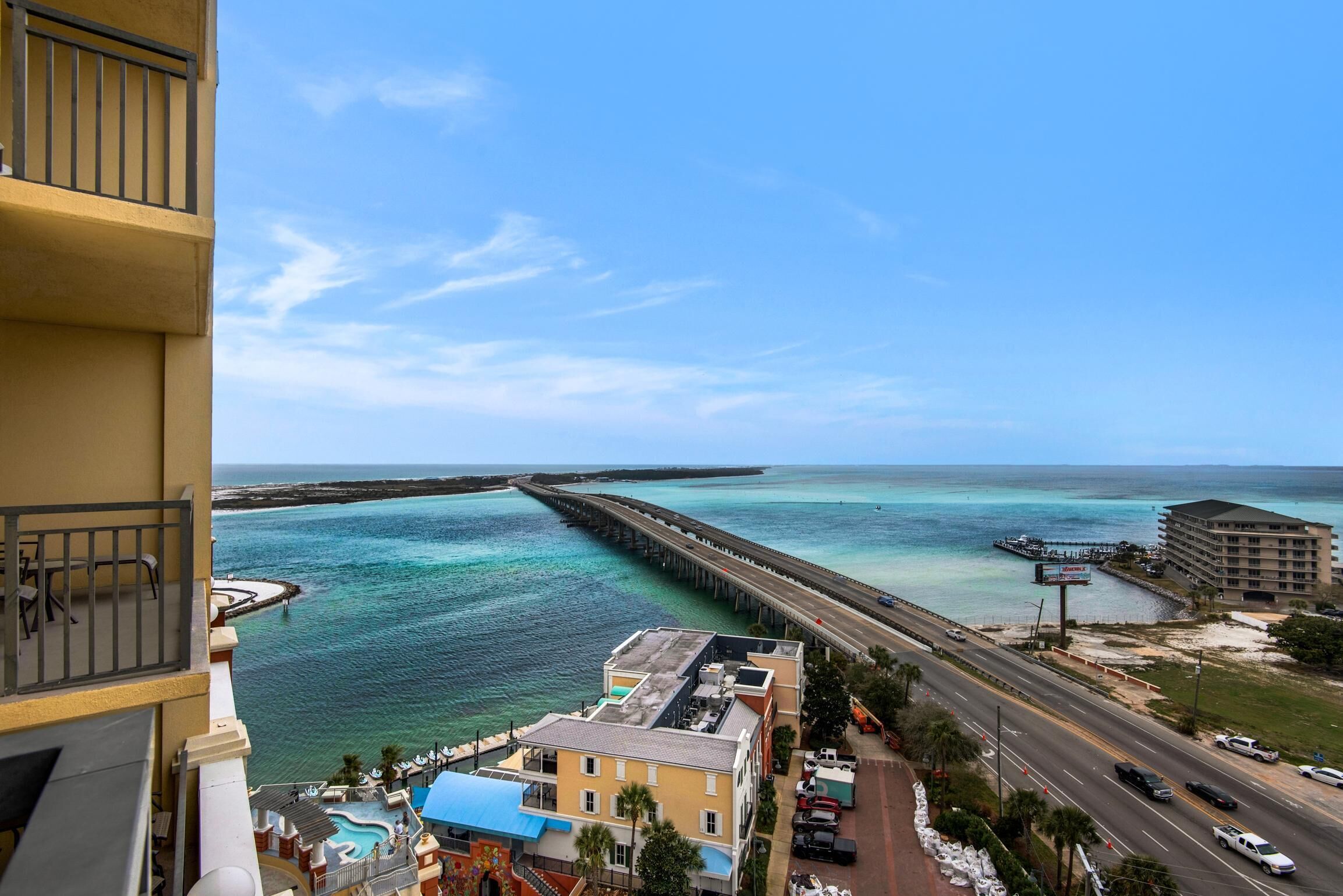 A coastal view with a pier extending into turquoise waters, a road with vehicles, and buildings including a hotel with a pool area.