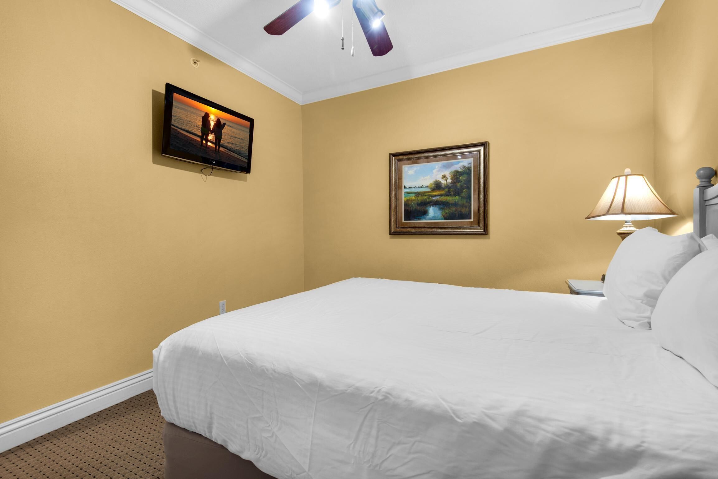 A hotel room with a large bed, a television mounted on the wall, a framed picture, a nightstand with a lamp, and a ceiling fan.