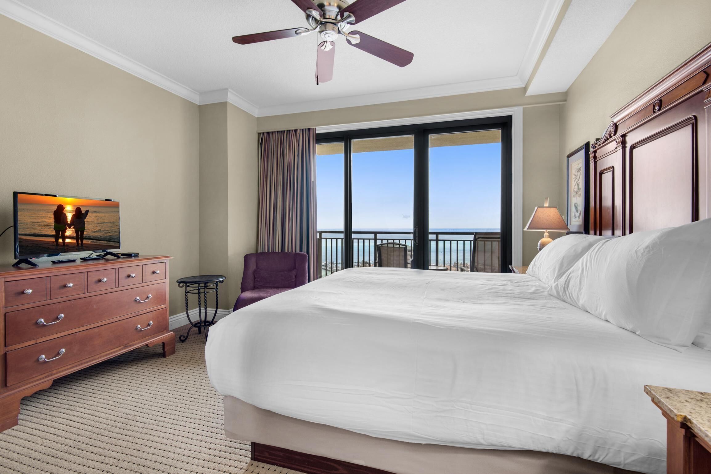 Un cuarto de hotel con una cama grande, un ventilador de techo, un televisor sobre un aparador de madera, una silla y un balcón con vistas al cielo.