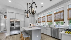 A modern kitchen with a central island, stainless steel appliances, and a large window with bamboo blinds. - Cypress by the Sea (Santa Rosa Beach)