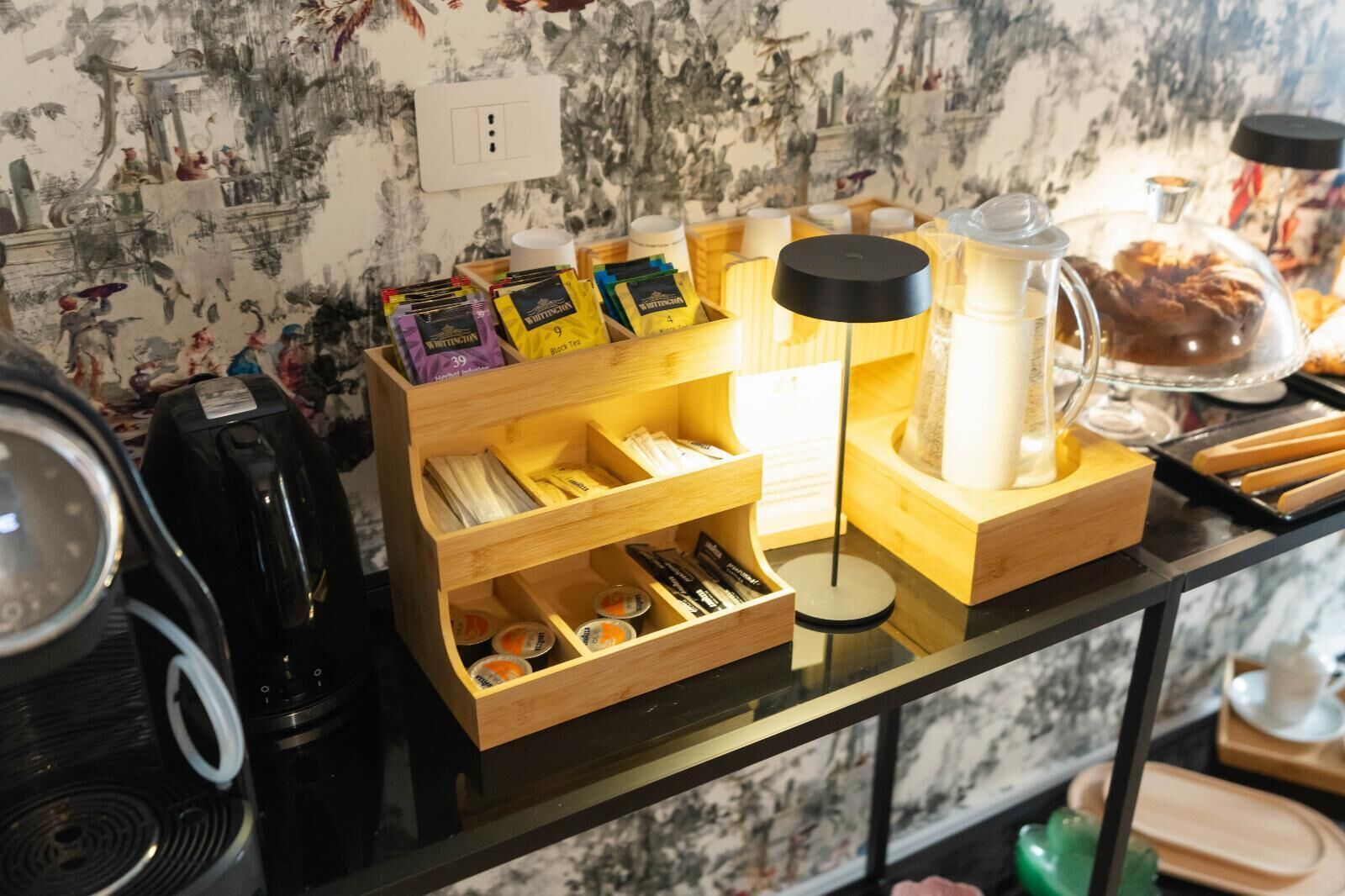 A coffee station with various tea and coffee products, a coffee maker, and a glass container with a cake.