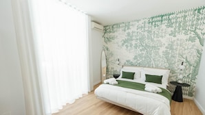 A bedroom with a bed, a bedside table, a wall-mounted air conditioner, and a window with sheer curtains.
