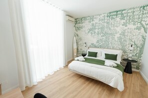 Un dormitorio con cama, mesita de noche, aire acondicionado instalado en la pared y ventana con cortinas de gasa.