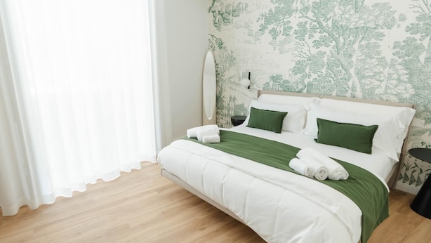 A neatly made bed with white and green bedding, a bedside table with a lamp, and a wall with a nature-themed wallpaper.