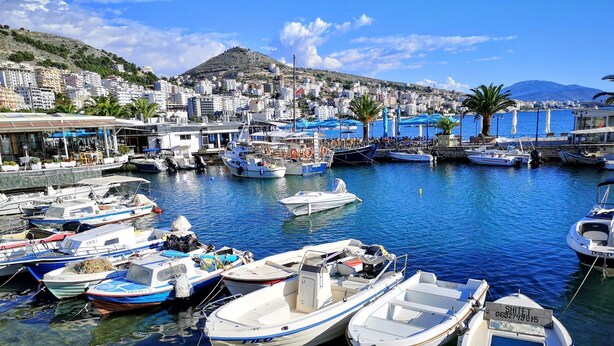 A marina with various boats docked, a clear blue sky, and a hilly backdrop with buildings.