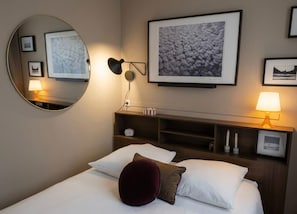 Een hotelkamer met een bed, een ronde spiegel, een wandplank met een klok, ingelijste foto's en een nachtkastje met een lamp.