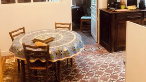 Un coin repas avec une table ronde recouverte d’une nappe à motifs floraux, entourée de chaises en bois. Le sol est décoré de carreaux formant un motif.
