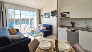A modern living room with a sofa, dining table set for two, and a kitchen area with a microwave and dishwasher.