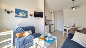 A modern living room with a blue sofa, a dining table set for two, a kitchen area, and a television mounted on the wall.