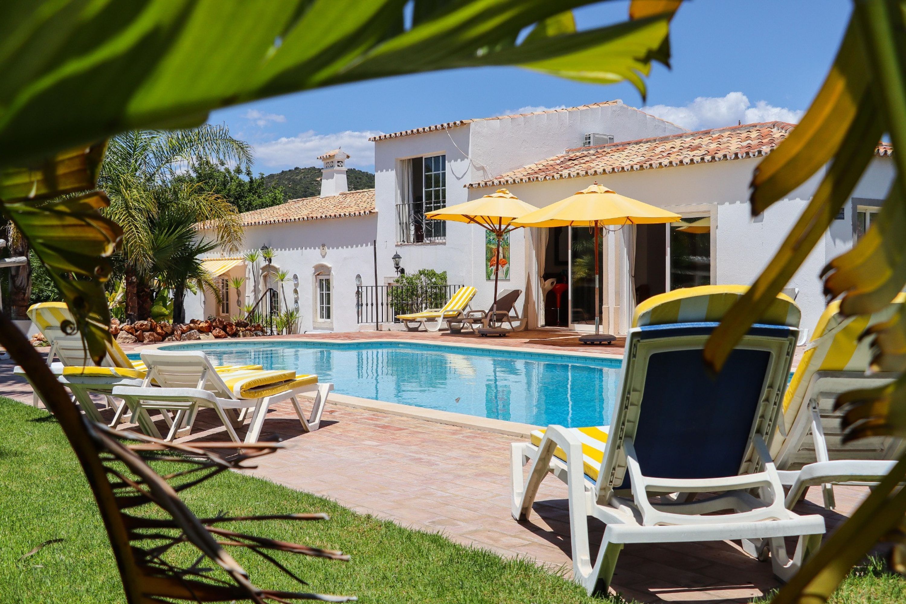A pool area with lounge chairs and umbrellas, a white building with a tiled roof, and a clear sky.