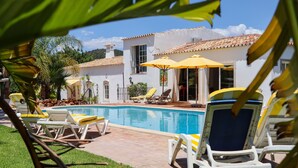 A pool area with lounge chairs and umbrellas, a white building with a tiled roof, and a clear sky.