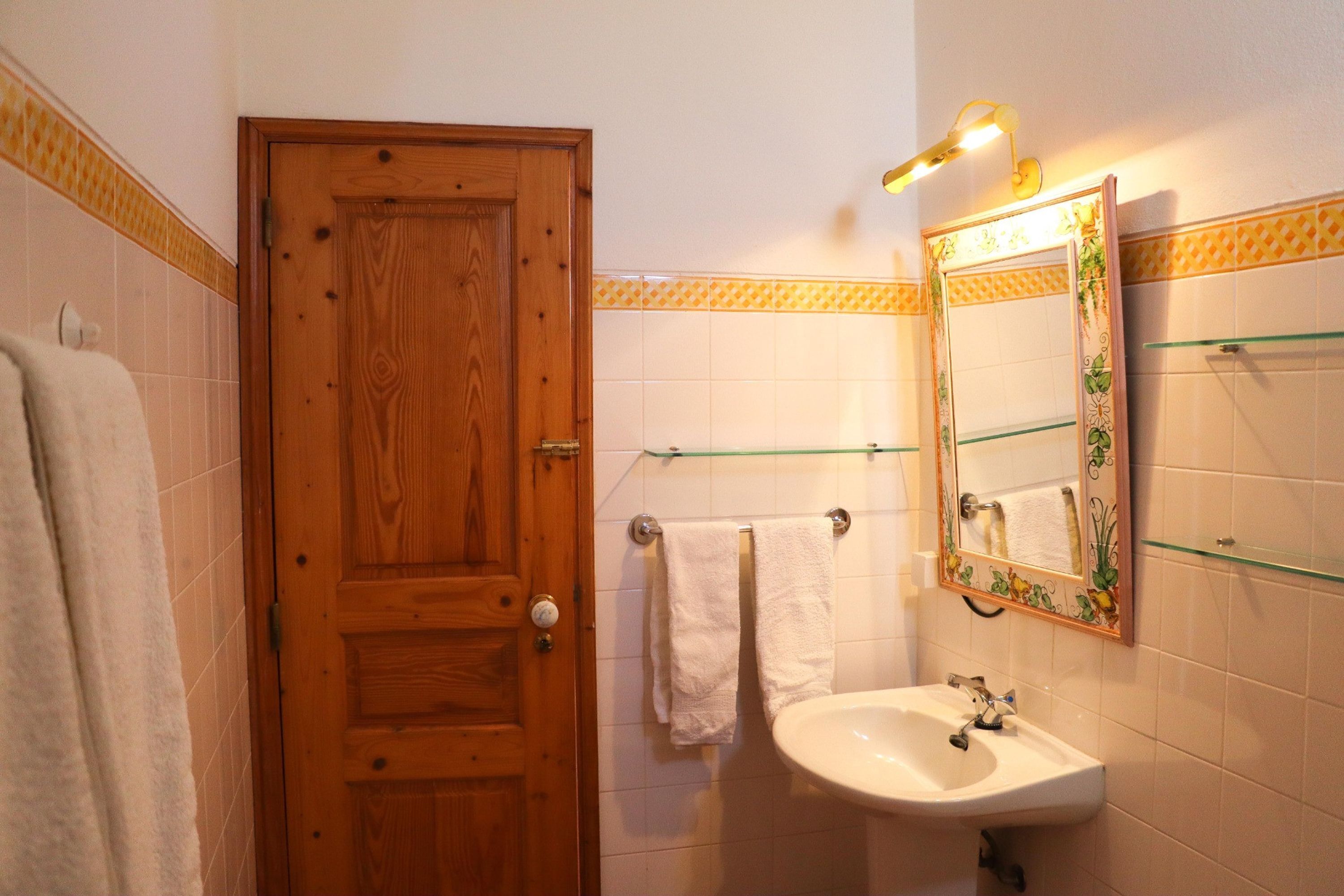 A bathroom with a wooden door, white tiled walls, a glass shelf, a sink, and a mirror with a decorative frame.