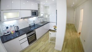Uma cozinha moderna com armários brancos, bancada preta e micro-ondas. Há uma pia, um fogão e uma cadeira. O chão é de madeira e as paredes são brancas.