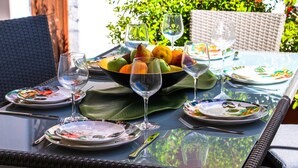 Mesa posta com pratos, talheres e uma cesta de frutas, com copos e uma cadeira de vime ao fundo.