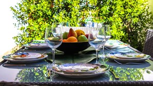 Mesa posta para refeição, com tigela de frutas e taças de vinho, cercada por pratos e talheres, com um denso hedge verde ao fundo.