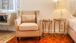 Um quarto com uma poltrona bege, uma mesa lateral redonda com abajur, uma cama e piso de madeira.