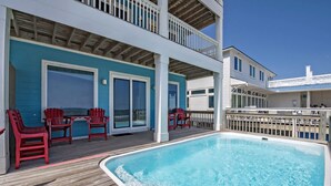 A pool area with a wooden deck, red Adirondack chairs, and a two-story building with a blue exterior.