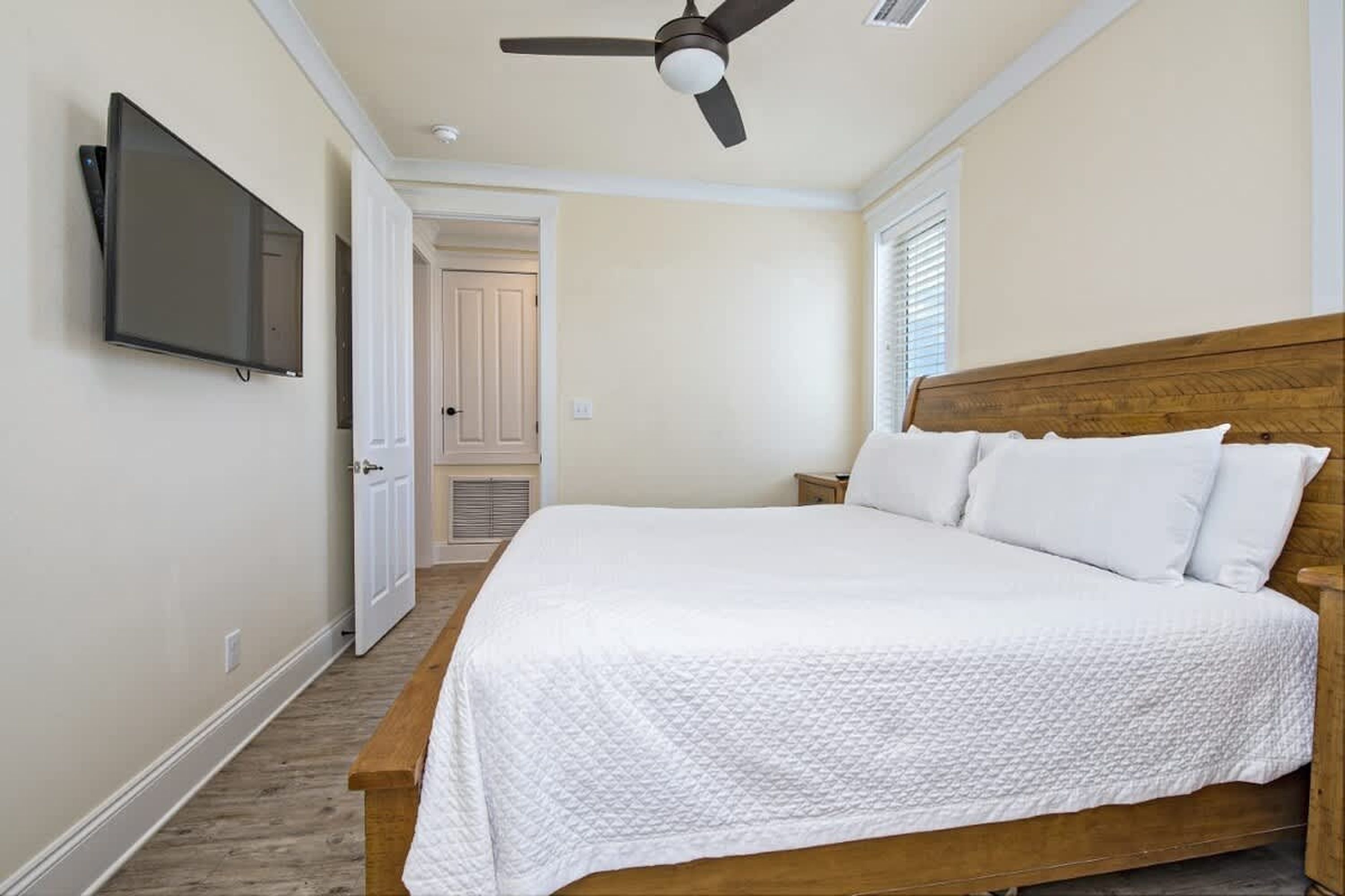 Un dormitorio con una cama, un ventilador de techo, un televisor montado en la pared y una puerta que da a otra habitación.