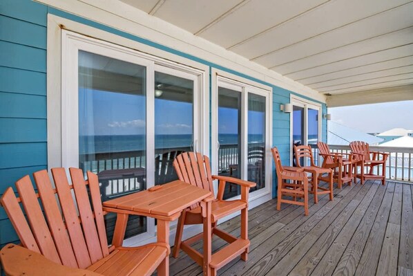 A covered deck with wooden chairs and tables, large sliding glass doors, and a view of the ocean. - Beachfront 9bed Easybreezy East (Gulf Shores)