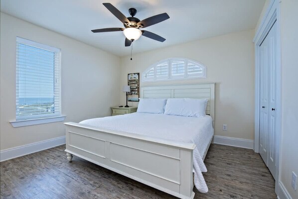 A bedroom with a white bed, a ceiling fan, a nightstand, and a window with blinds. - Beachfront 9bed Easybreezy East (Gulf Shores)