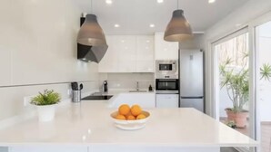 A modern kitchen with white cabinets, a central island with a bowl of oranges, and pendant lighting. - Villas2meet Casa Blanca Porto Cristo (Porto Cristo)