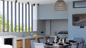 A modern kitchen with a large island, a built-in grill, and a dining area set for six. - Villa Vacasa - A Heaven of Luxury in the Heart of Crete (Aggeliana)