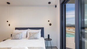 A modern bedroom with a large bed, two bedside tables, and a view of a pool and mountains through a glass door. - Villa Vacasa - A Heaven of Luxury in the Heart of Crete (Aggeliana)